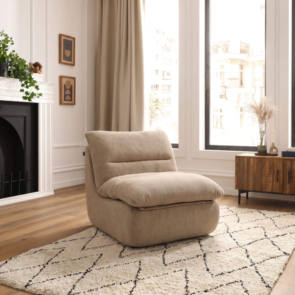 Un fauteuil beige confortable installé dans un salon lumineux avec parquet, tapis géométrique, cheminée et grandes fenêtres créant une ambiance chaleureuse et moderne.