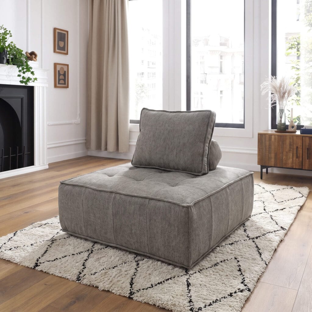 Un fauteuil bas modulable gris en tissu avec coussin d'appoint, posé sur un tapis berbère dans un salon lumineux aux tons naturels et chaleureux.