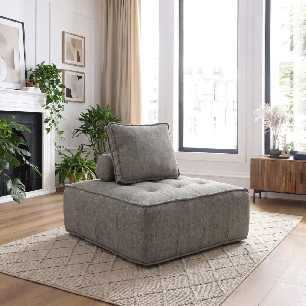 Un fauteuil-pouf gris capitonné dans un salon lumineux au style contemporain, avec plantes vertes, cheminée et grandes fenêtres créant une ambiance apaisante